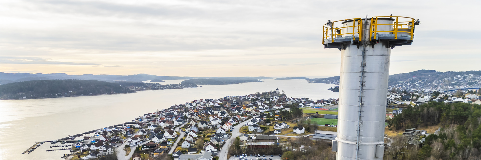 Utsikt over et kystområde med bolighus som strekker seg langs vannet til venstre. Til høyre står et høyt, hvitt tårn med en gul sikkerhetsplattform øverst. Skog og åser er synlige i bakgrunnen under en lys, overskyet himmel.