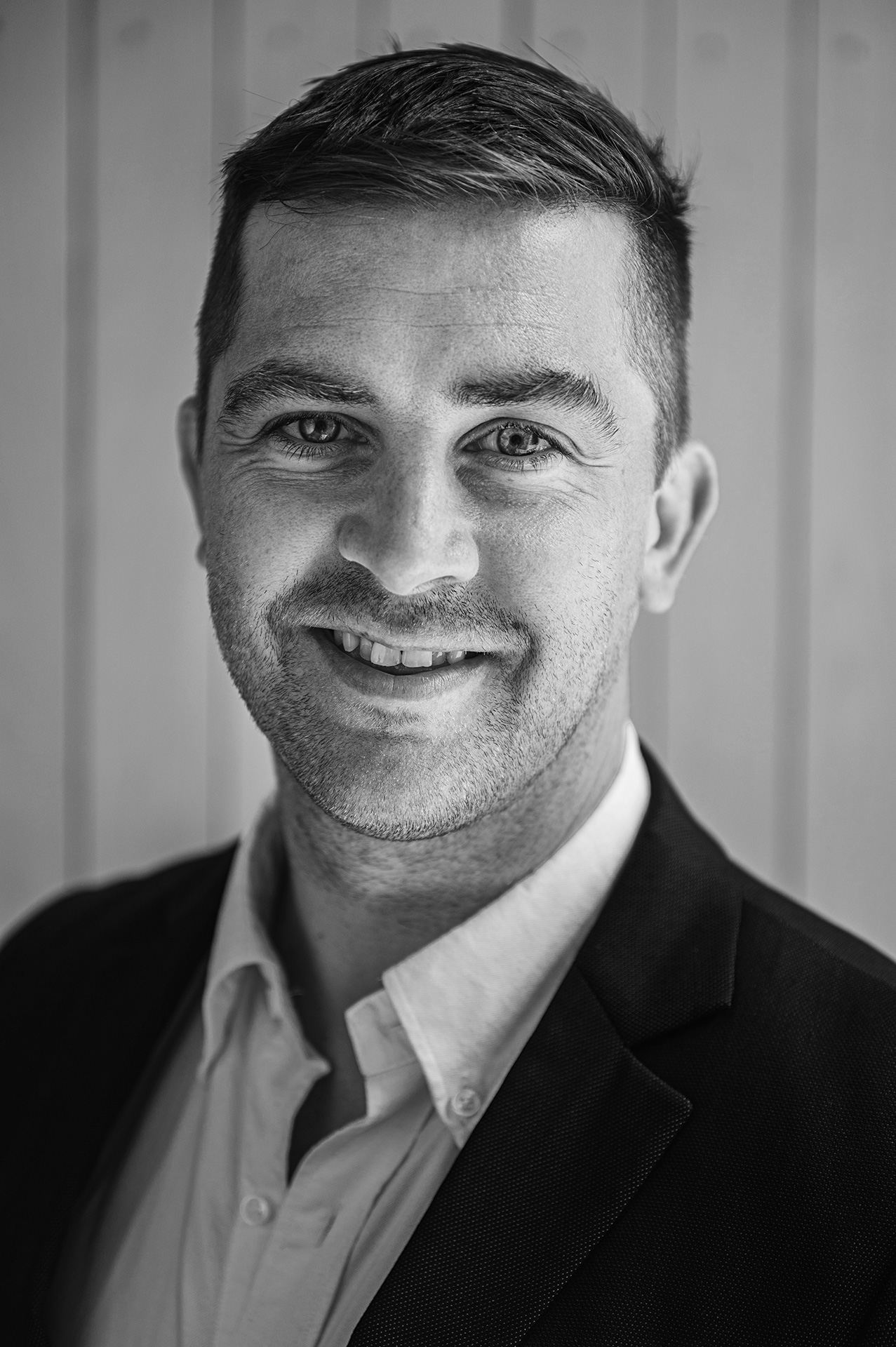 A man wearing a dark blazer and light dress shirt, standing against a vertical wooden panel background.