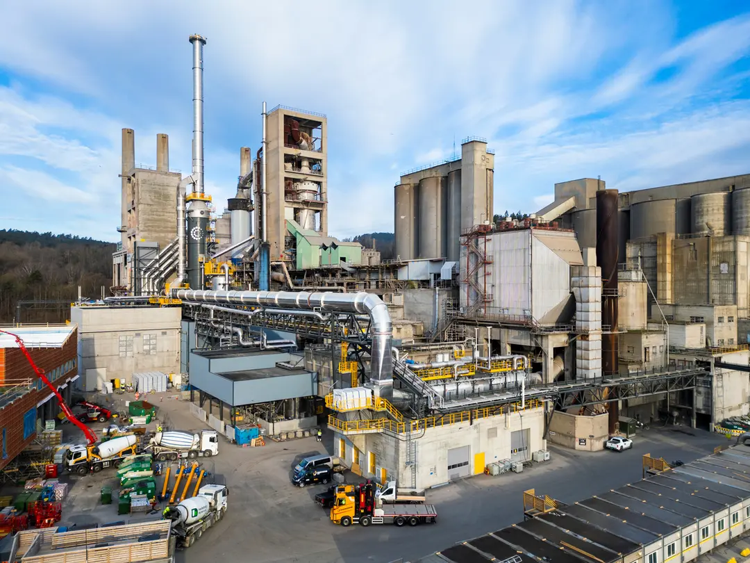 Industrial cement factory with large interconnected buildings, silos, and smokestacks under a blue sky. Machinery, vehicles, and equipment are visible in the foreground, with surrounding greenery in the distance.