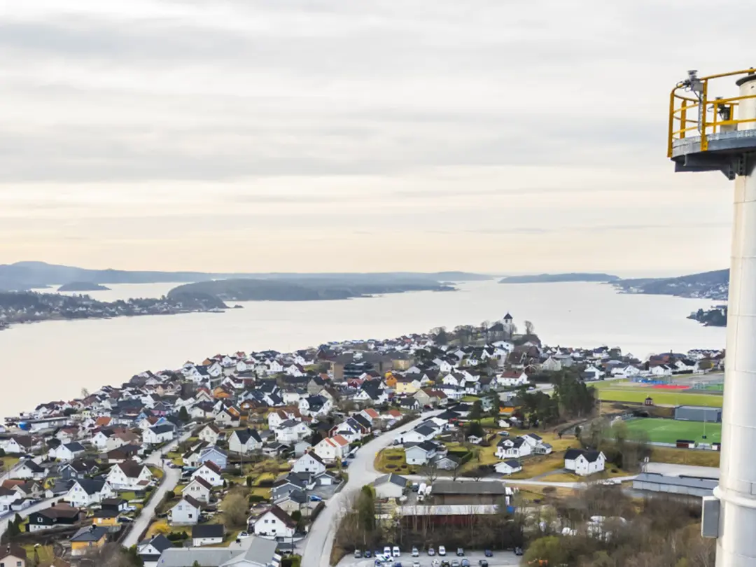 Utsikt over et kystområde med bolighus som strekker seg langs vannet til venstre. Til høyre står et høyt, hvitt tårn med en gul sikkerhetsplattform øverst. Skog og åser er synlige i bakgrunnen under en lys, overskyet himmel.