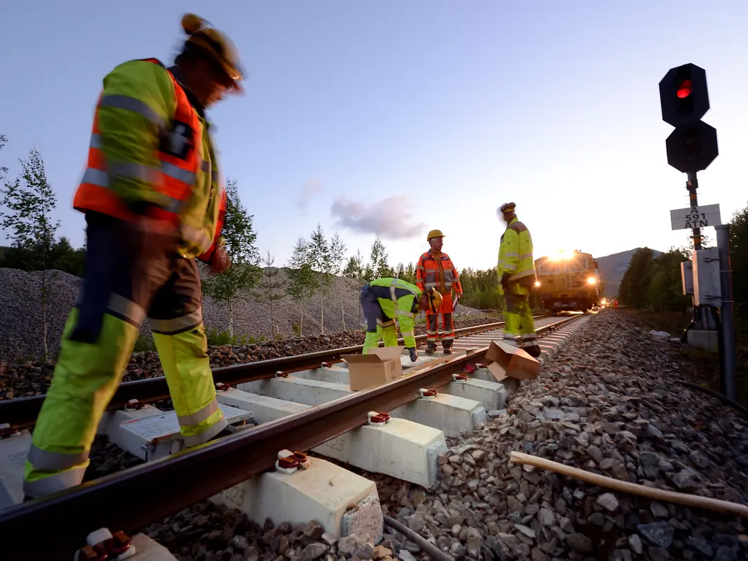 Arbetare i reflexkläder reparerar ett järnvägsspår i skymningen, med ett rött stoppljus till höger och skog i bakgrunden.